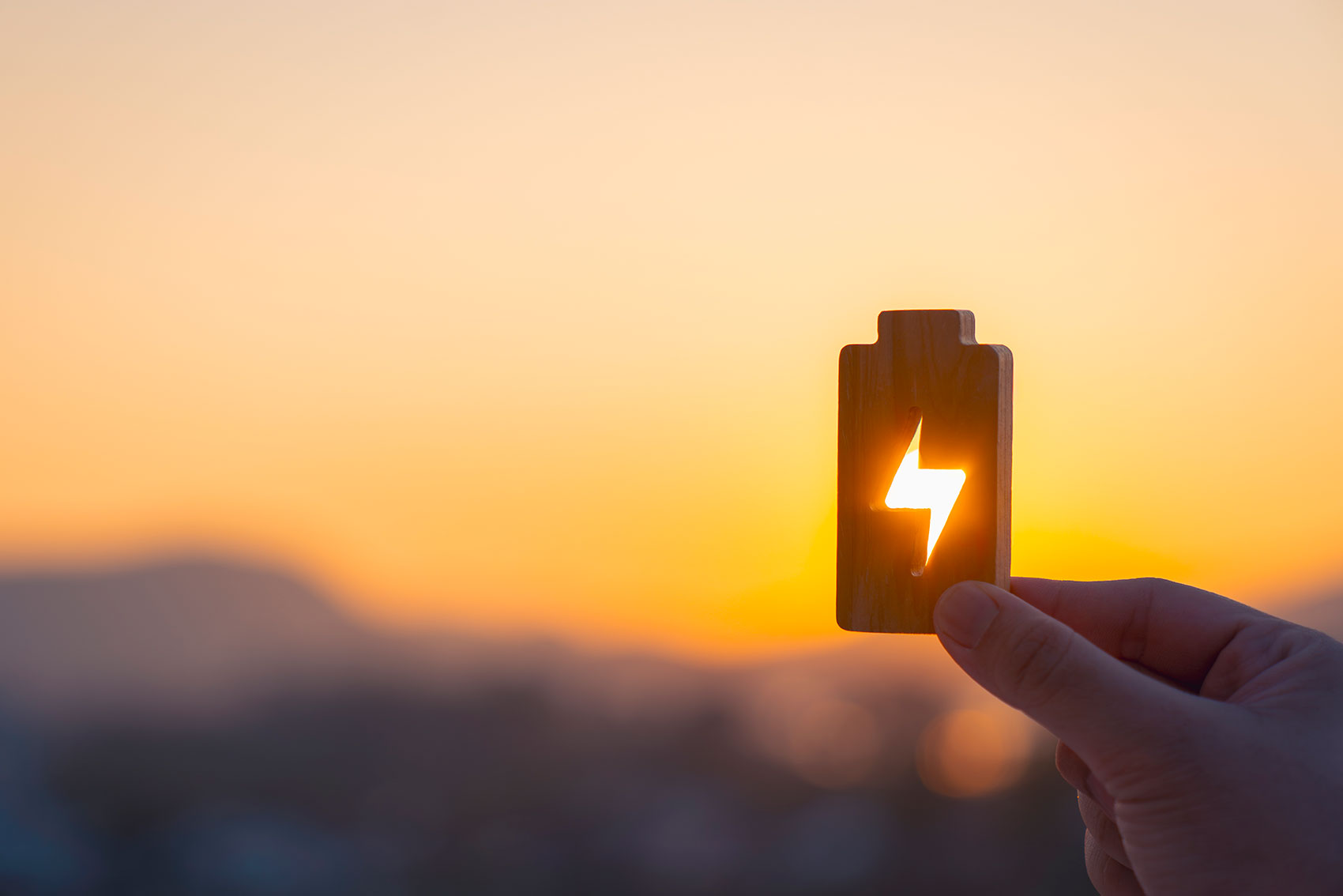 Hand holding a glowing battery icon against a sunrise